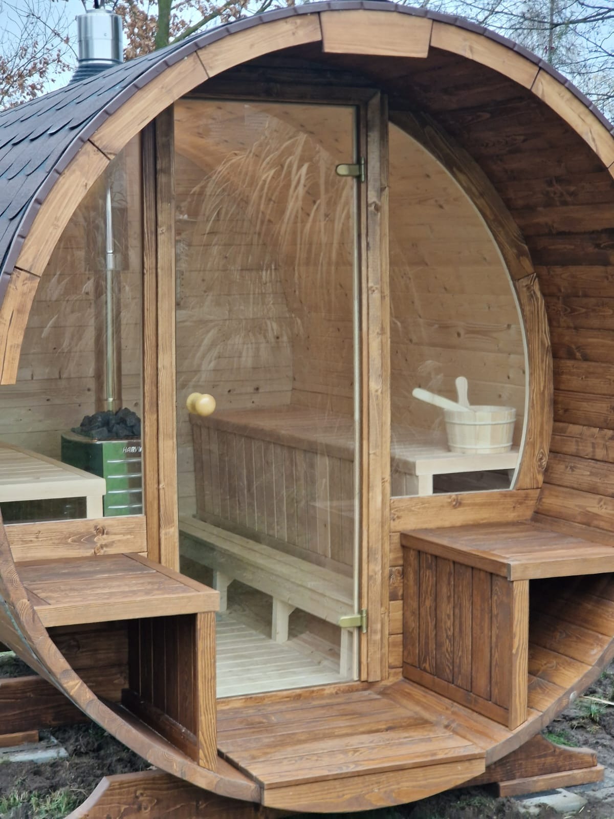 Drewniana sauna w kształcie beczki z przeszklonymi drzwiami, widoczny piec i akcesoria do sauny. Całość w ciepłych odcieniach brązu, na zewnątrz.