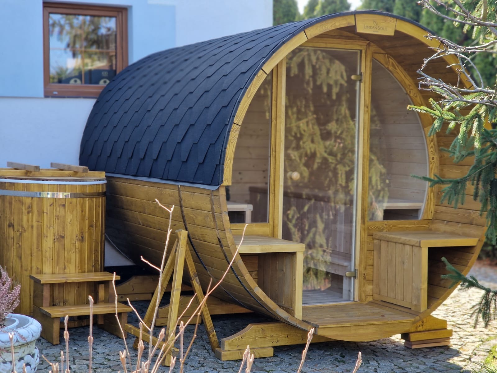 Drewniana sauna beczka z czarnym dachem i przeszklonymi drzwiami, obok drewniana balia. Całość na tle domu i zieleni. Widoczne schodki i podpory sauny.