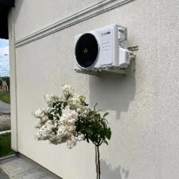 Biała jednostka zewnętrzna klimatyzacji Bosch zamontowana na elewacji budynku, z kwitnącym krzewem hortensji poniżej i widocznym logo firmy Cool House Service w rogu.