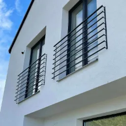 Biały budynek z dwoma małymi balkonami z czarnymi, prostymi barierkami, widok z dołu na tle błękitnego nieba z chmurami.