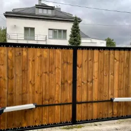 Dwuskrzydłowa brama wjazdowa z drewna o ciepłym odcieniu, osadzona w czarnej metalowej ramie z ozdobnymi nitami, wyposażona w automatyczny system otwierania, na tle domu z szarą dachówką.
