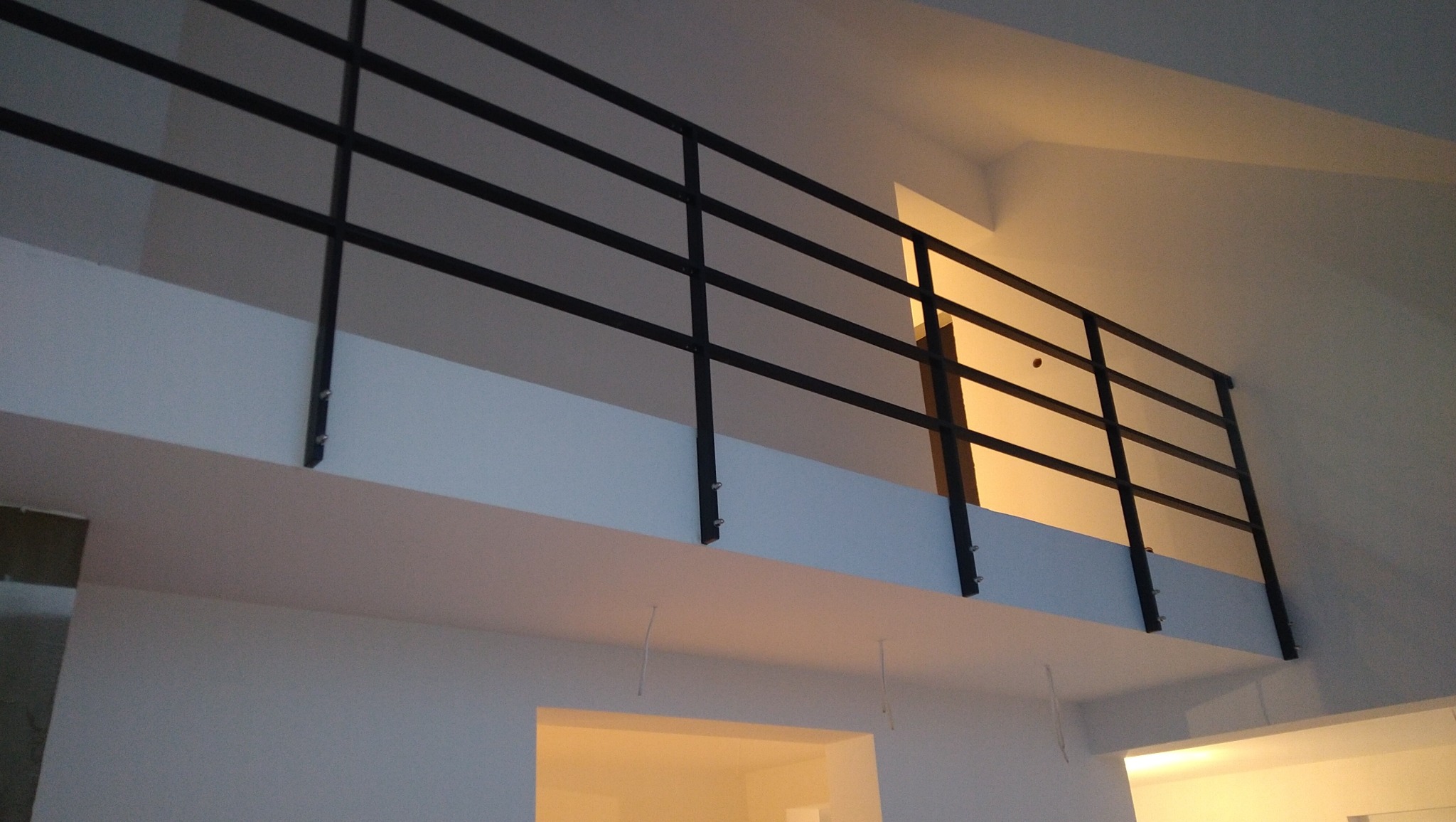 Minimalistyczna czarna balustrada metalowa z czterema poziomymi prętami, zamontowana na białej antresoli z widocznymi śrubami mocującymi, oświetlona ciepłym światłem zza ściany.