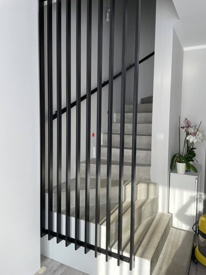 Nowoczesne schody z szarego betonu z czarną, pionową konstrukcją metalową pełniącą funkcję balustrady i dekoracyjnego elementu oddzielającego przestrzeń.