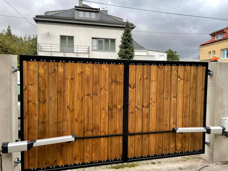 Dwuskrzydłowa brama wjazdowa z drewna o ciepłym odcieniu, osadzona w czarnej metalowej ramie z ozdobnymi nitami, wyposażona w automatyczny system otwierania, na tle domu z szarą dachówką.