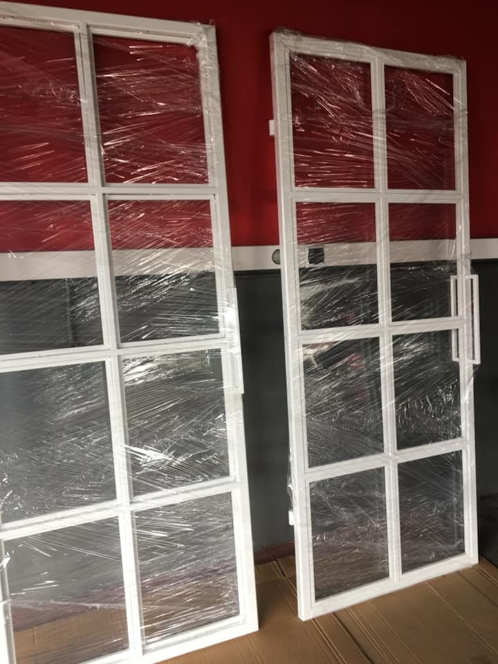 Dwa białe, metalowe panele z kwadratowymi podziałami, owinięte folią ochronną, opierają się o ścianę z czerwonym i szarym pasem. Jeden panel ma zamontowany uchwyt.