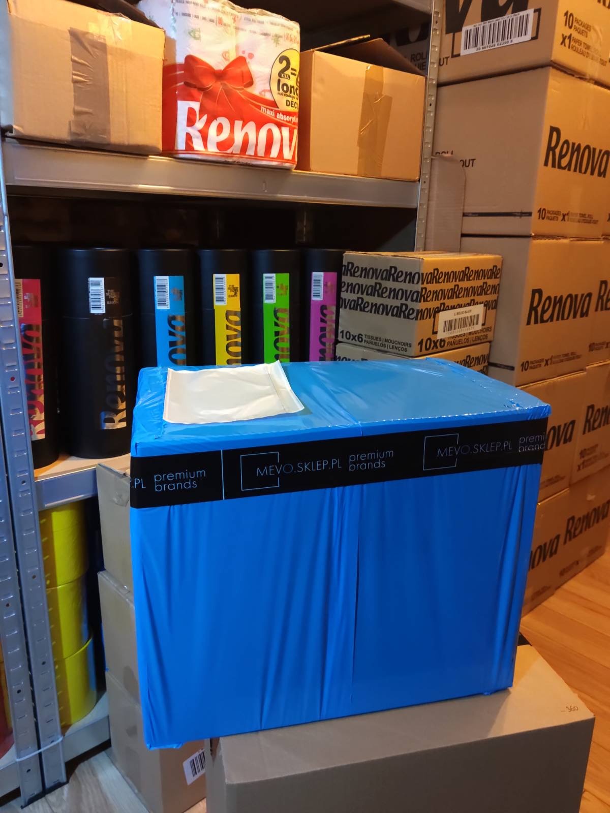 Magazyn z zapasem artykułów higienicznych Renova, w tle czarne pojemniki z kolorowymi etykietami i kartony z napisem Renova, na pierwszym planie niebieska paczka z czarnym paskiem z napisem mevo.