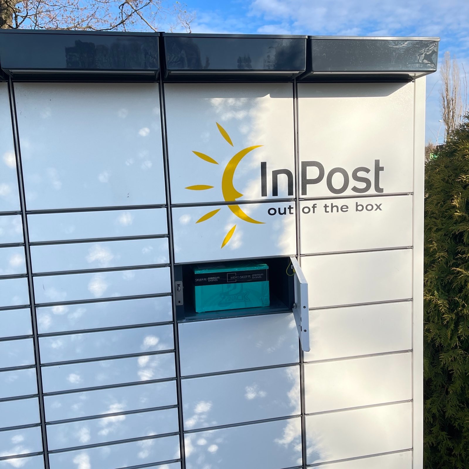 Paczkomat InPost z otwartą skrytką, wewnątrz widoczne turkusowe pudełko z produktami, na zewnątrz odbicie błękitnego nieba.
