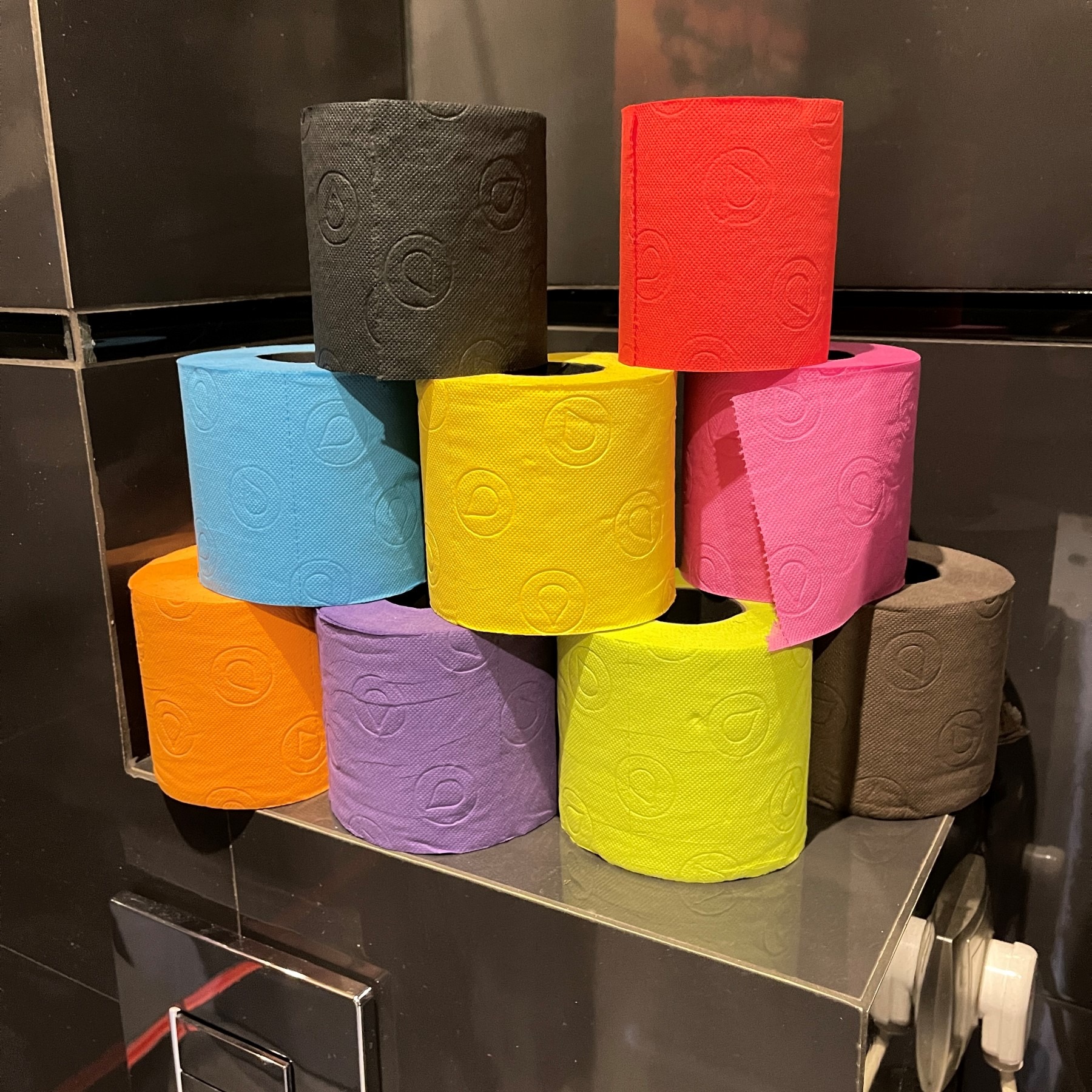 Stos kolorowych rolek papieru toaletowego (czarny, czerwony, niebieski, żółty, pomarańczowy, fioletowy, limonkowy, brązowy) z wytłoczonym wzorem, ułożony na czarnej, błyszczącej półce w łazience.