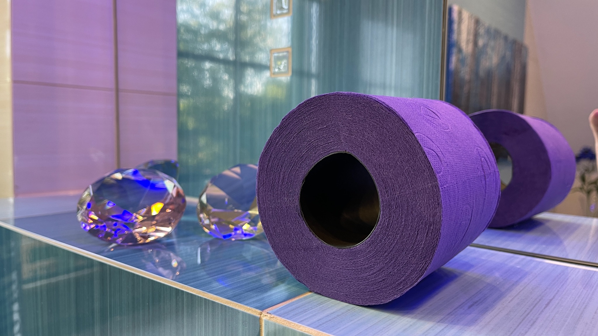 Dwa rolki fioletowego papieru toaletowego i dekoracyjne szklane diamenty na niebieskiej, błyszczącej półce w łazience w Bielsku-Białej, odbijające się w lustrze.