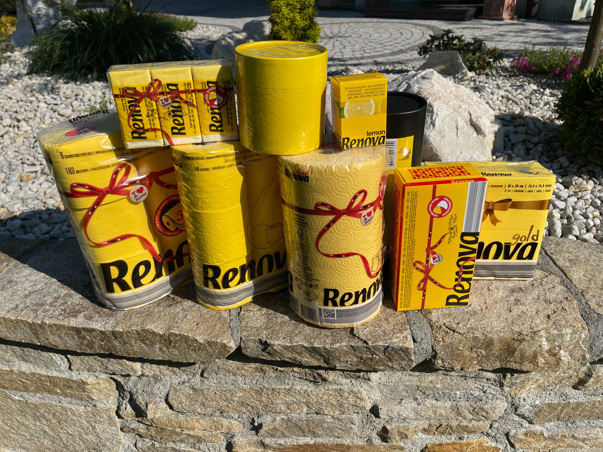 Żółte rolki papieru toaletowego i chusteczki odświeżające marki Renova, ułożone na kamiennym murku na zewnątrz, w słoneczny dzień.