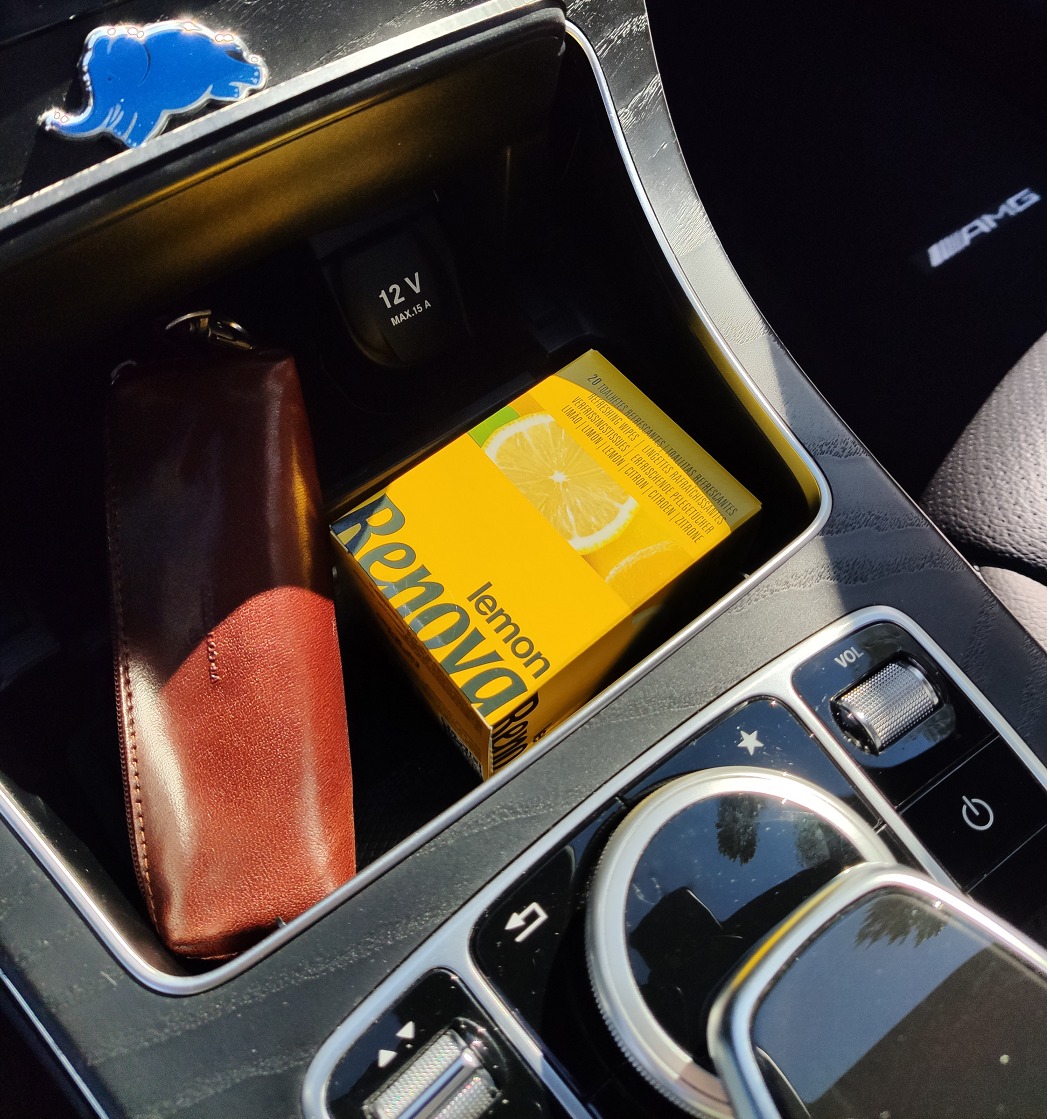 Żółte opakowanie nawilżanych chusteczek Renova Lemon w schowku samochodowym Mercedes-Benz AMG, obok brązowego etui na klucze i gniazda 12V.