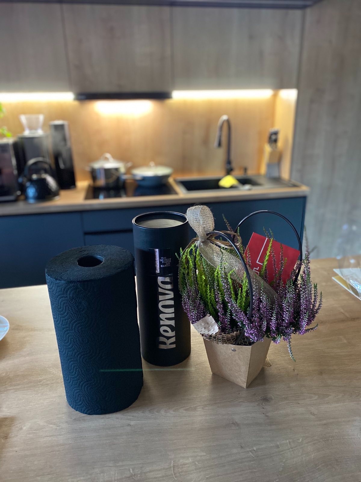 Czarny ręcznik papierowy Renova, pojemnik z napisem Renova oraz wrzos w ozdobnym koszyczku na drewnianym blacie kuchennym; w tle widoczny zlewozmywak i kuchenka.