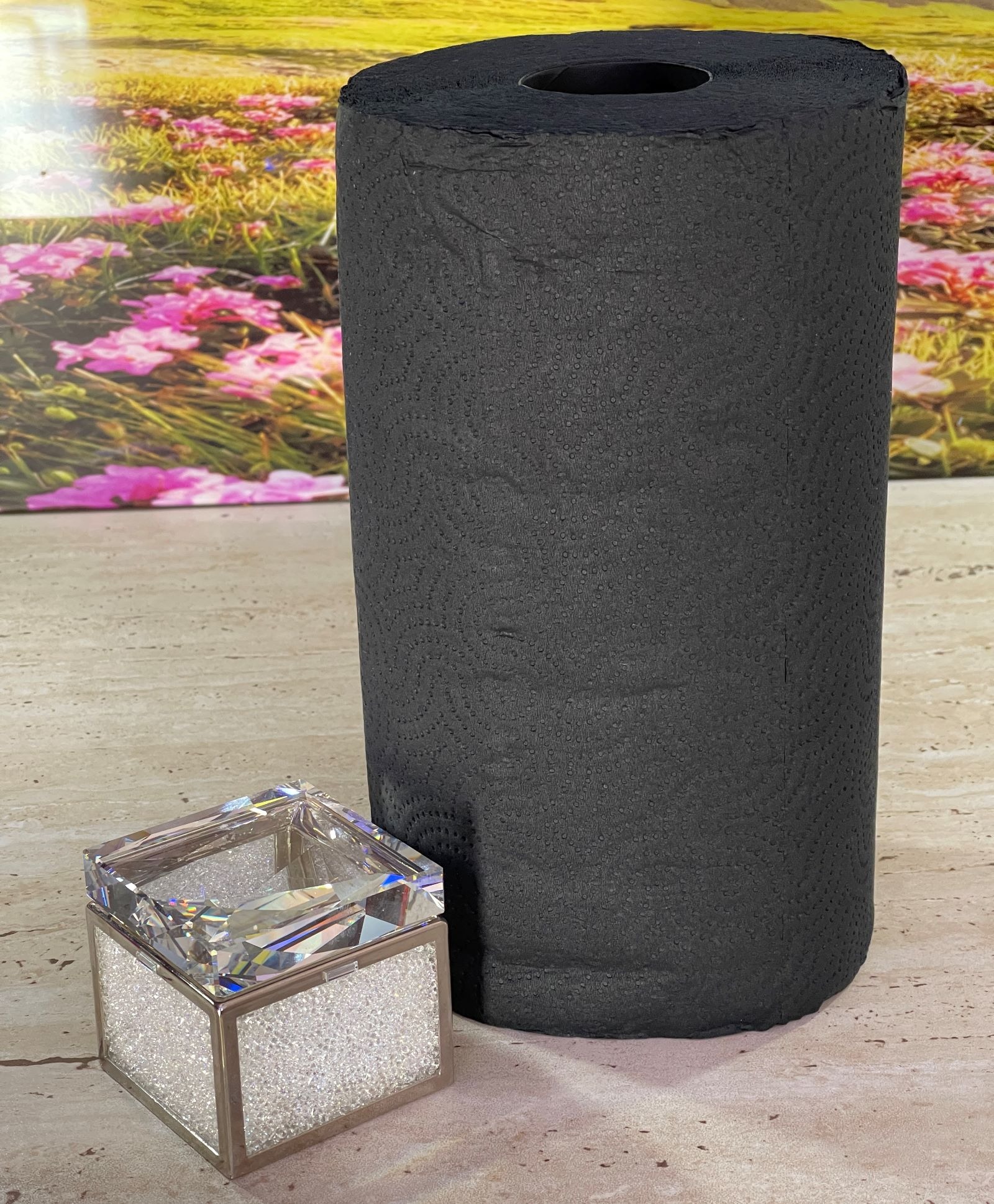 Rolka czarnych ręczników papierowych obok dekoracyjnego pudełka z kryształkami na tle rozmytej łąki z kwiatami.