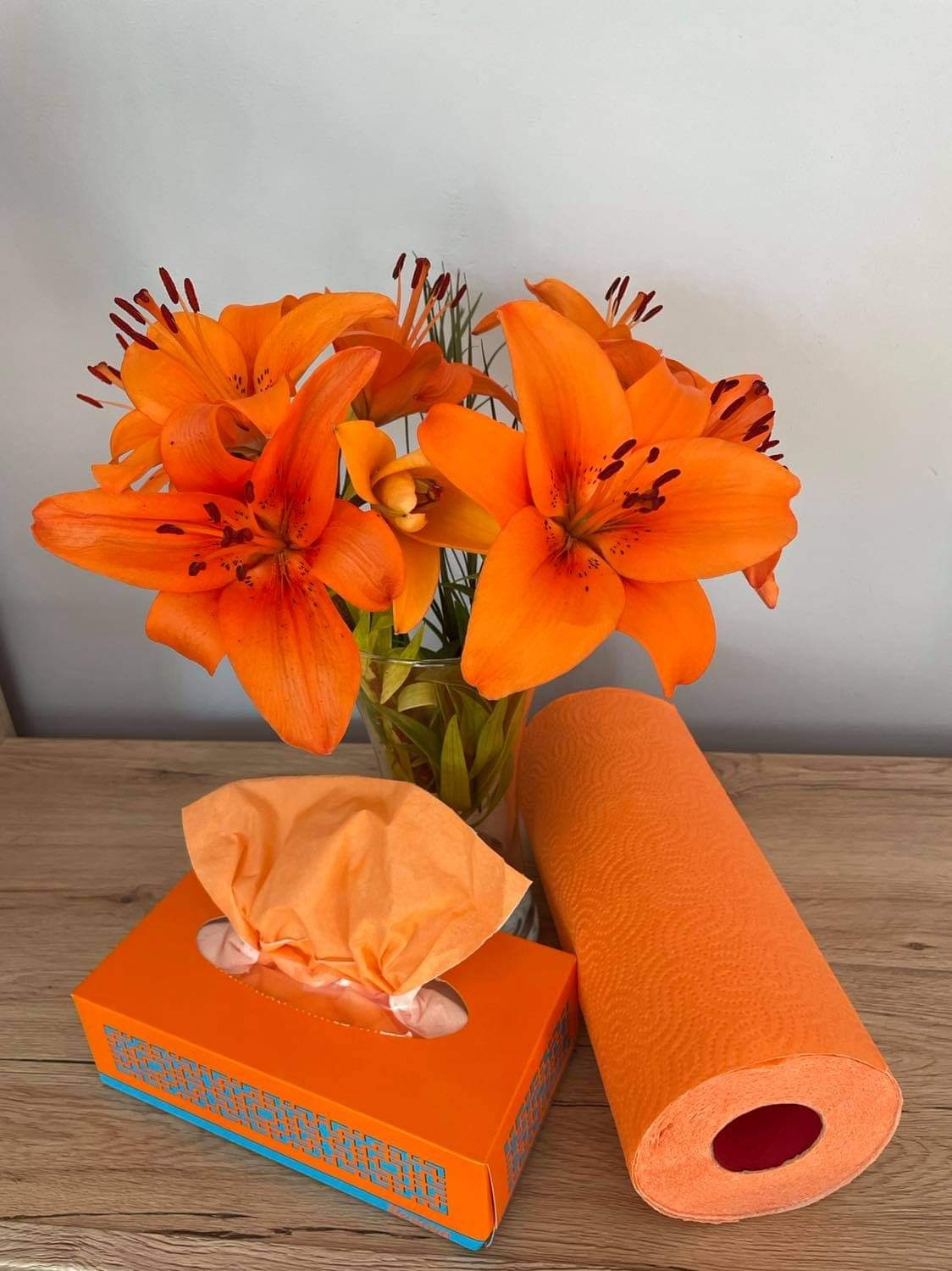 Kompozycja z pomarańczowych lilii w wazonie, pudełka chusteczek higienicznych i rolki ręcznika papierowego w odcieniach pomarańczy na drewnianym blacie.