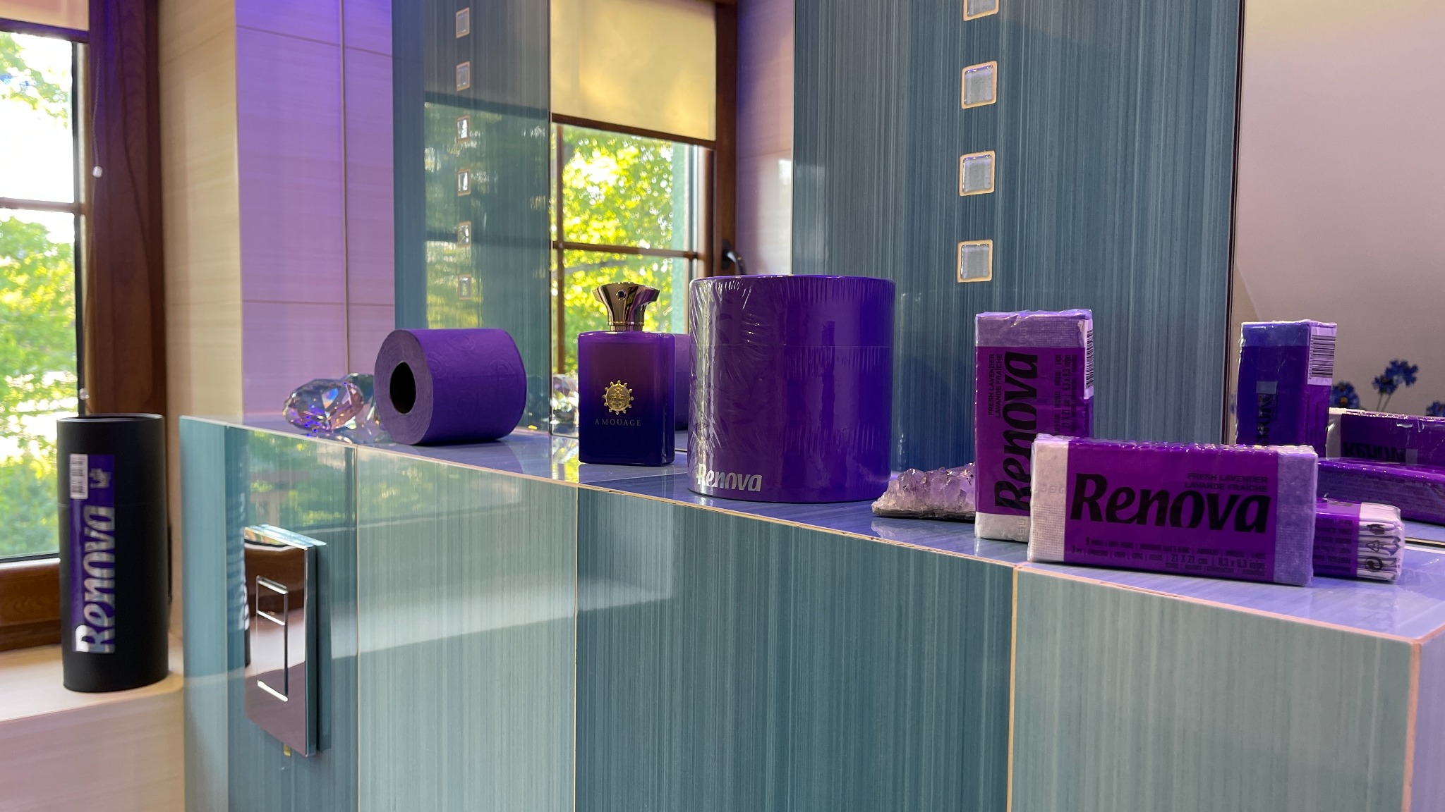Fioletowe akcesoria łazienkowe Renova, w tym papier toaletowy, ręczniki papierowe i pudełko, ustawione na niebieskiej płytce w łazience z oknem i flakonem perfum Amouage.
