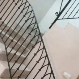 Klatka schodowa z czarną, metalową balustradą o dekoracyjnym wzorze, widok z dołu na schody z jasnymi stopniami i ścianą w odcieniu beżu.