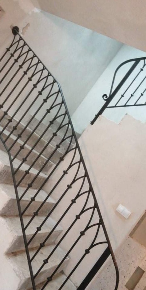 Klatka schodowa z czarną, metalową balustradą o dekoracyjnym wzorze, widok z dołu na schody z jasnymi stopniami i ścianą w odcieniu beżu.