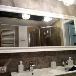 Nowoczesna łazienka z dwoma umywalkami, dużym lustrem w białej ramie i widoczną kabiną prysznicową w odbiciu. Ciemna, marmurowa ściana dodaje elegancji.