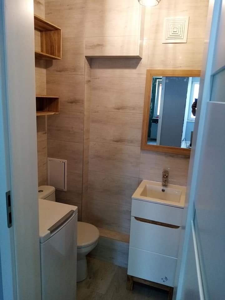 Ukończona łazienka z beżowymi płytkami imitującymi drewno, wisząca szafka z umywalką, toaleta z pralką obok, drewniane półki na ścianie, lustro w drewnianej ramie i sufitowa lampa.