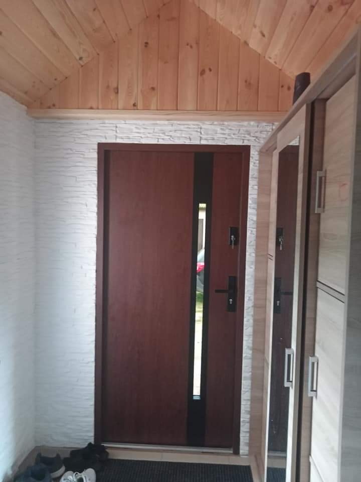 Wnętrze przedpokoju z drewnianym sufitem, ścianą wyłożoną białą cegłą dekoracyjną, brązowymi drzwiami z wąskim przeszkleniem i częścią szafy z lustrem