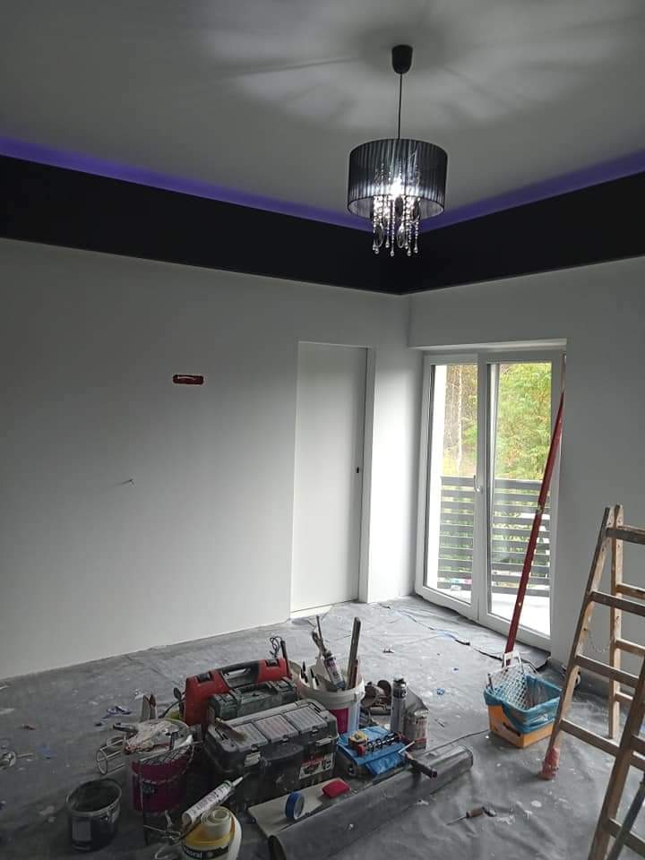 Pokój w trakcie prac wykończeniowych: świeżo pomalowane białe ściany, czarny pas pod sufitem z fioletowym podświetleniem LED, czarny żyrandol z kryształkami, podłoga zabezpieczona folią, narzędzia...
