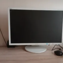 Biały monitor NEC z czarnym ekranem na biurku z myszką i plątaniną kabli, oświetlony z boku.