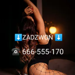 Kobieta w czarnym body i kabaretkach, leżąca na łóżku w ciemnym pomieszczeniu z numerem telefonu wyświetlonym na zdjęciu.