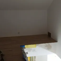 Fragment wnętrza w trakcie układania paneli podłogowych, widoczne narzędzia, fragment drabiny i zabezpieczona taśmą malarską ściana.