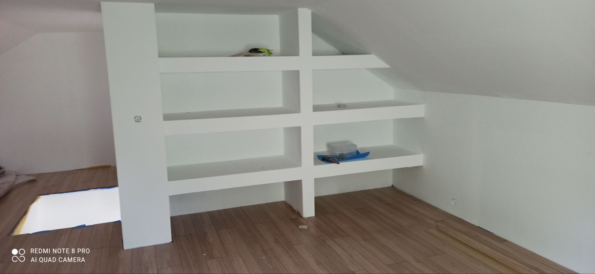 Półki z płyty karton gips wnękowe w skosie poddasza, z widocznymi narzędziami i pojemnikiem na śruby na jednej z półek, podłoga wyłożona panelami.