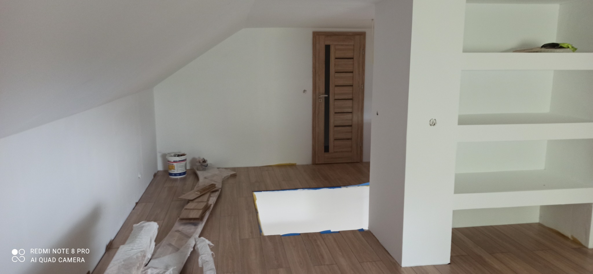 Poddasze po remoncie: świeżo pomalowane białe ściany, drewniana podłoga w trakcie układania, biała zabudowa regałowa, drzwi wewnętrzne z przeszkleniami, wiadro z farbą w rogu.