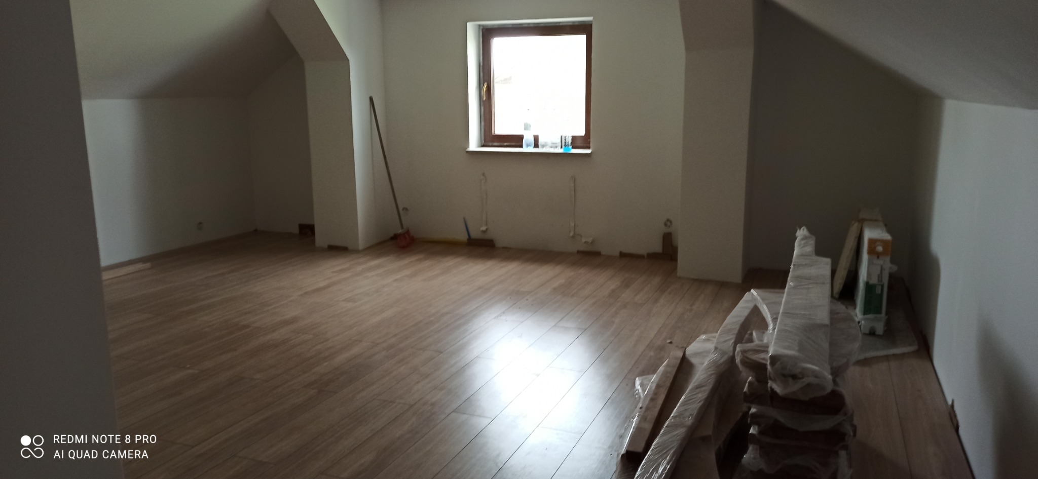 Poddasze po remoncie z nową drewnianą podłogą, panele w folii czekają na montaż, widoczna niedokończona listwa przypodłogowa i ślady po instalacjach na ścianie.