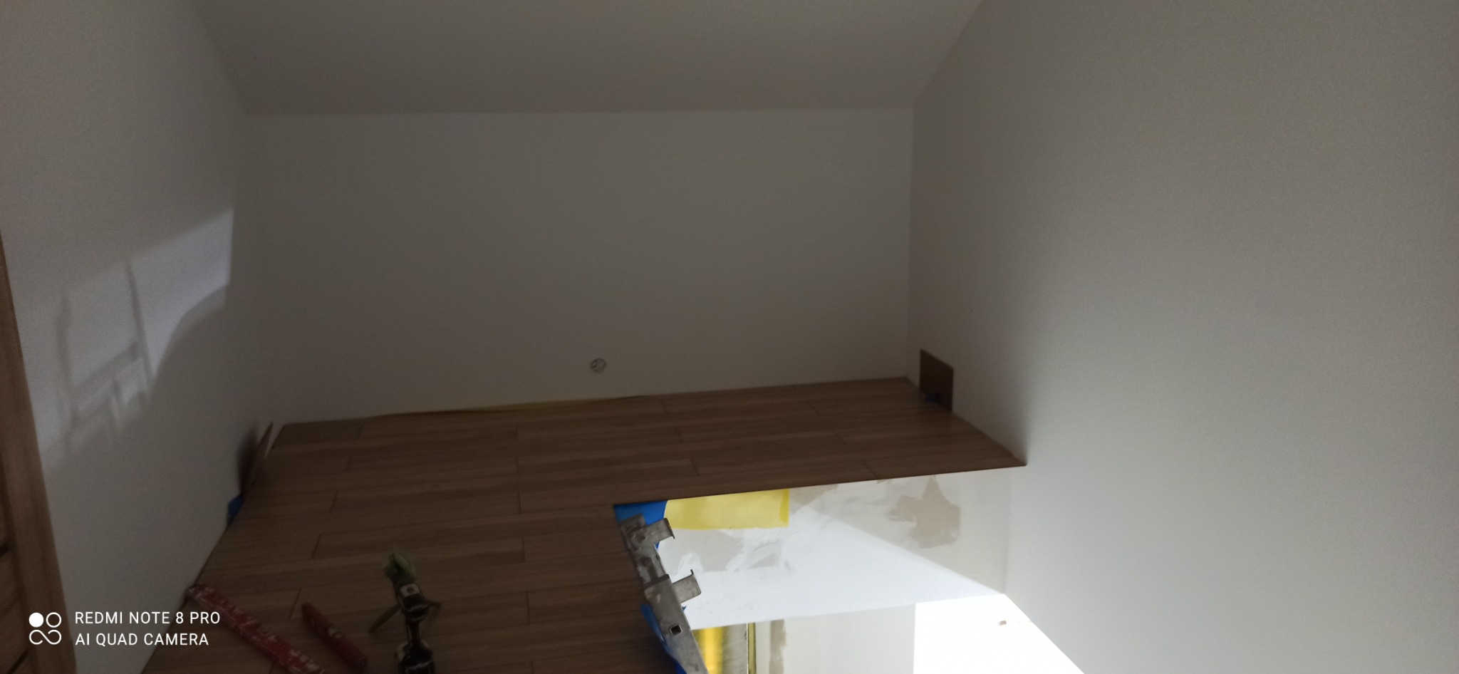 Fragment wnętrza w trakcie układania paneli podłogowych, widoczne narzędzia, fragment drabiny i zabezpieczona taśmą malarską ściana.