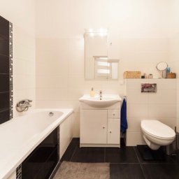 Marek Waszczuk - Wykończona łazienka w bieli i czerni, z białą wanną, umywalką z szafką, toaletą i ciemną podłogą z płytek. Widoczny fragment lustra i akcesoria łazienkowe na półce.