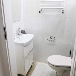 Marek Waszczuk - Biała, minimalistyczna łazienka z umywalką nablatową, szafką podumywalkową z chromowanymi uchwytami, toaletą i grzejnikiem drabinkowym na tle białych płytek ceramicznych.