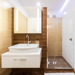 Marek Waszczuk - Nowoczesna łazienka z umywalką nablatową, lustrem z podświetleniem LED i kabiną prysznicową ze szklanymi drzwiami, wykończona płytkami w odcieniach beżu i brązu, z ciemną drewnopodobną podłogą...