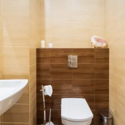 Nowoczesna łazienka z beżowo-brązowymi płytkami, wiszącą toaletą, chromowanym koszem na śmieci i dekoracyjną muszlą na półce.