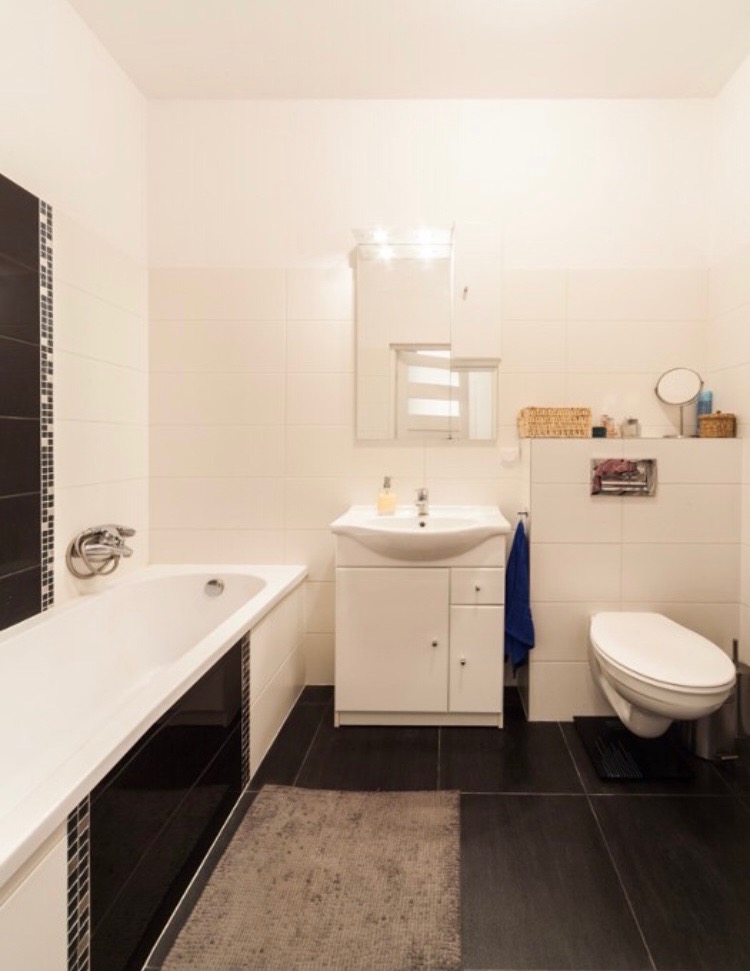Wykończona łazienka w bieli i czerni, z białą wanną, umywalką z szafką, toaletą i ciemną podłogą z płytek. Widoczny fragment lustra i akcesoria łazienkowe na półce.