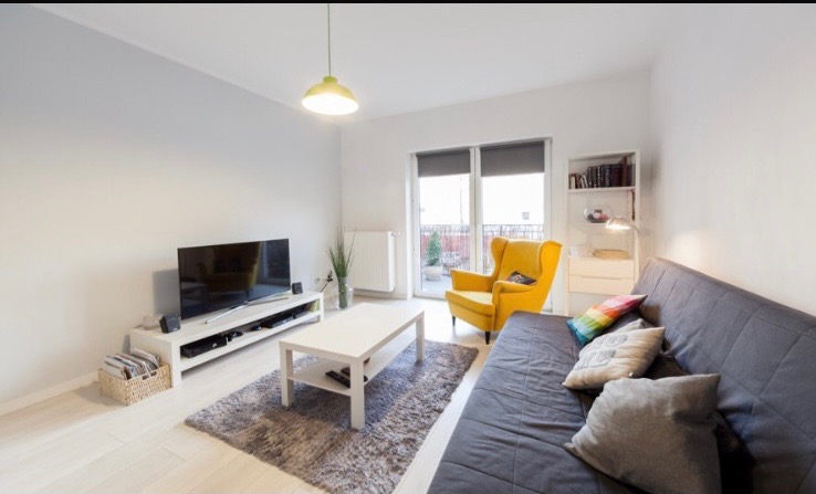 Jasne wnętrze salonu z drewnianą podłogą, szarą sofą, żółtym fotelem typu uszak i białymi ścianami, balkon z widokiem na zieleń w tle.