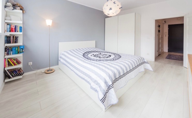 Jasna sypialnia z białymi panelami podłogowymi, szarą ścianą i białym łóżkiem z narzutą w biało-szare pasy oraz lampą podłogową i półką z książkami.