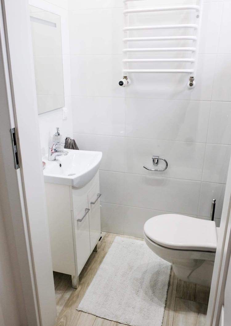 Biała, minimalistyczna łazienka z umywalką nablatową, szafką podumywalkową z chromowanymi uchwytami, toaletą i grzejnikiem drabinkowym na tle białych płytek ceramicznych.