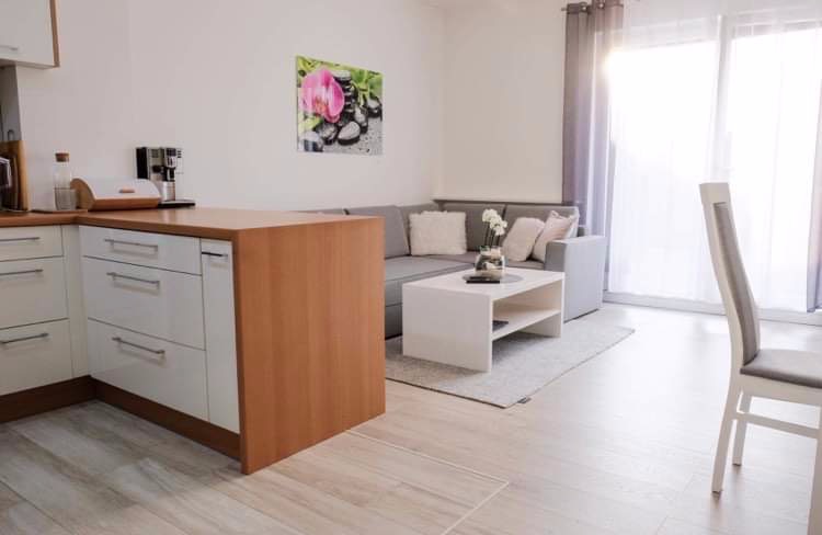 Jasne wnętrze mieszkania z drewnianą podłogą, szarą sofą, białym stolikiem kawowym i elementami zabudowy kuchennej w tle, oświetlone naturalnym światłem.