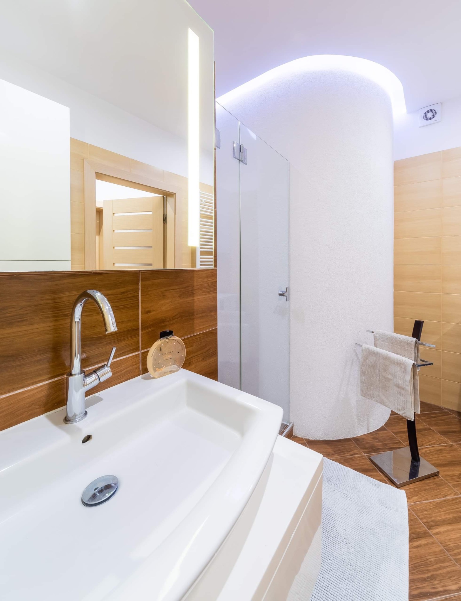 Nowoczesna łazienka z chromowaną armaturą, drewnopodobnymi płytkami ściennymi i podłogowymi, oraz kabiną prysznicową z podświetlanym sufitem.