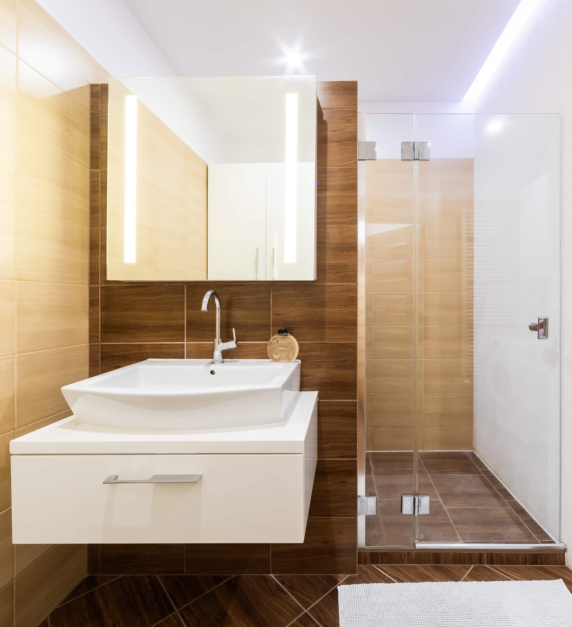 Nowoczesna łazienka z umywalką nablatową, lustrem z podświetleniem LED i kabiną prysznicową ze szklanymi drzwiami, wykończona płytkami w odcieniach beżu i brązu, z ciemną drewnopodobną podłogą...