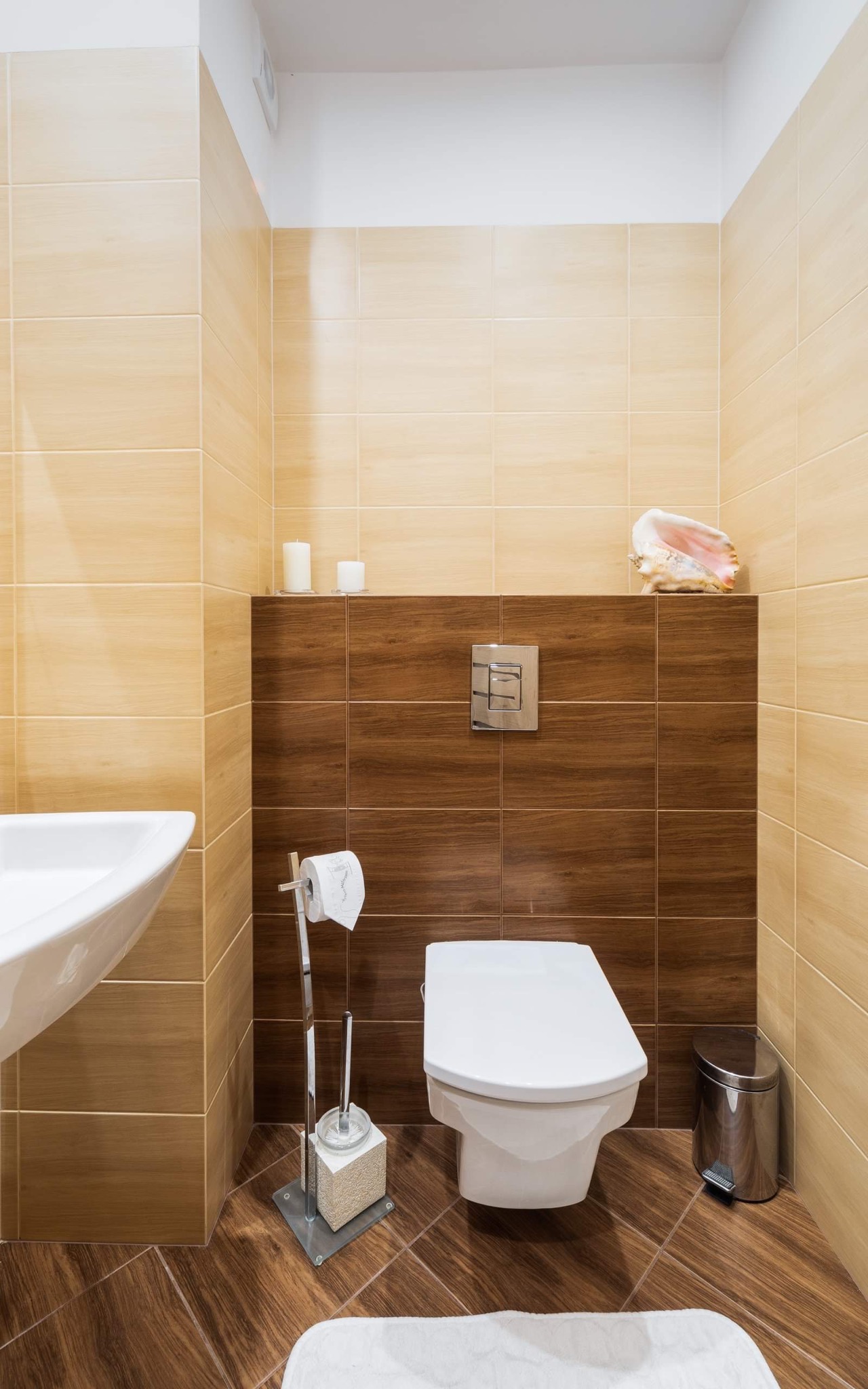 Nowoczesna łazienka z beżowo-brązowymi płytkami, wiszącą toaletą, chromowanym koszem na śmieci i dekoracyjną muszlą na półce.