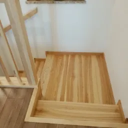 Fragment drewnianych schodów z jasnego drewna, widok z góry, z widoczną balustradą i dwoma stopniami, na tle drewnopodobnej podłogi i białej ściany z parapetem, na którym stoją doniczki z roślinami.