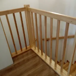 Fragment drewnianej balustrady przy schodach z widocznym oświetleniem ściennym i parkietem imitującym drewno.