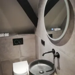 Nowoczesna łazienka na poddaszu z umywalką w kolorze antracytowym, okrągłym lustrem z podświetleniem LED i szarą płytką imitującą kamień.