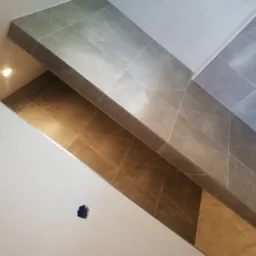 Wykończona łazienka z szaro-beżowymi płytkami imitującymi marmur, ułożonymi na ścianach i półce, widoczny otwór na gniazdko elektryczne w białej ścianie, oraz niebieska gąbka na podłodze.