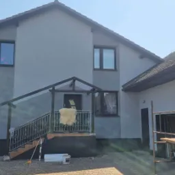 Dwukondygnacyjny dom z szarą elewacją po malowaniu, widoczne rusztowania i akcesoria malarskie, jasne okna z ciemnymi ramami, wejście z balustradą i dekoracyjnym futrem na poręczy.