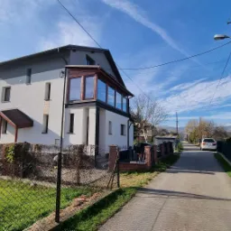 Dwukondygnacyjny dom jednorodzinny z dobudowanym przeszklonym balkonem, otynkowany na biało i szaro, widoczny z ulicy w słoneczny dzień.